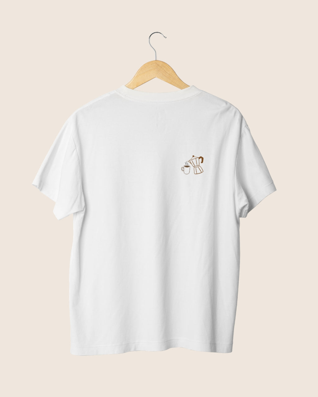 Detalle minimalista en el pecho de la camiseta blanca unisex Before Coffee, mostrando una ilustración lineal marrón de una cafetera moka y una taza humeante.